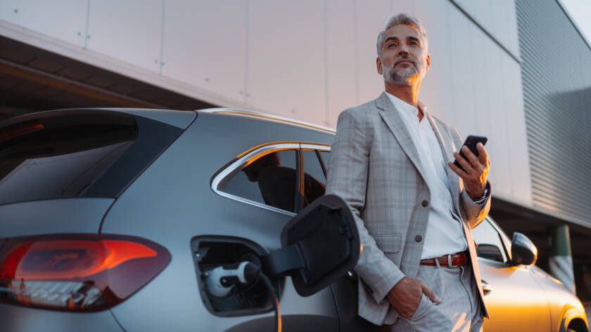 Businessman holding smartphone while charging car at electric vehicle charging station, closeup.