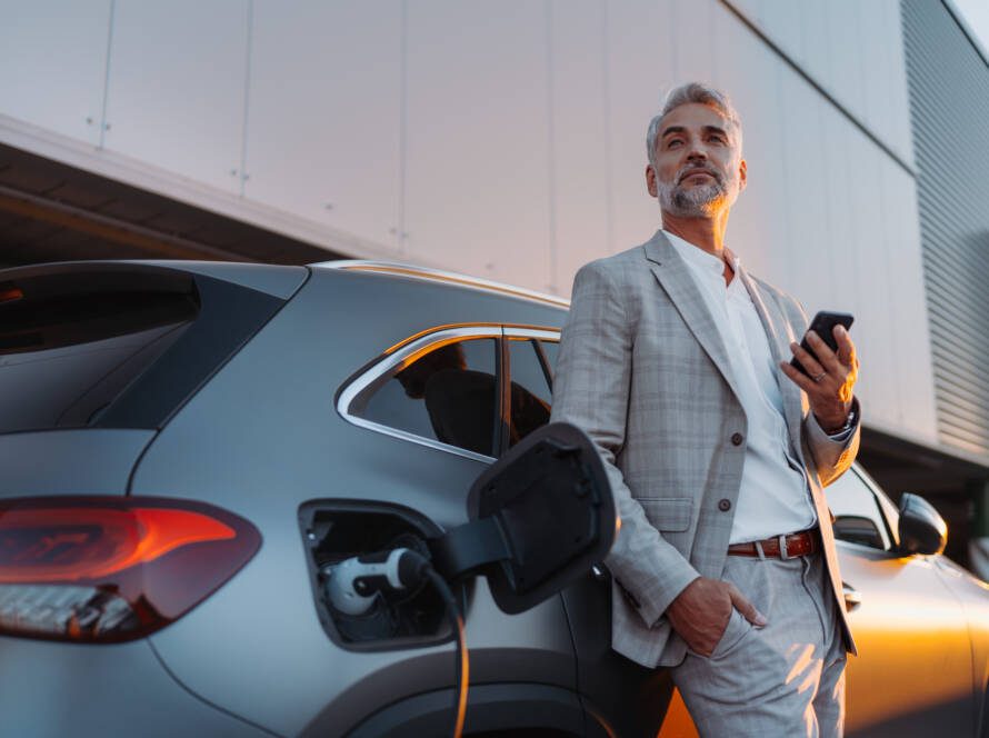 Businessman holding smartphone while charging car at electric vehicle charging station, closeup.